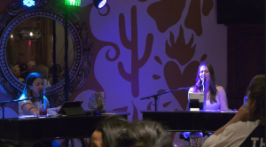 Kate Louise and her partner Laura play seperate pianos. the room features puprle lighting and the wall is shows a drawing of an orange cactus.